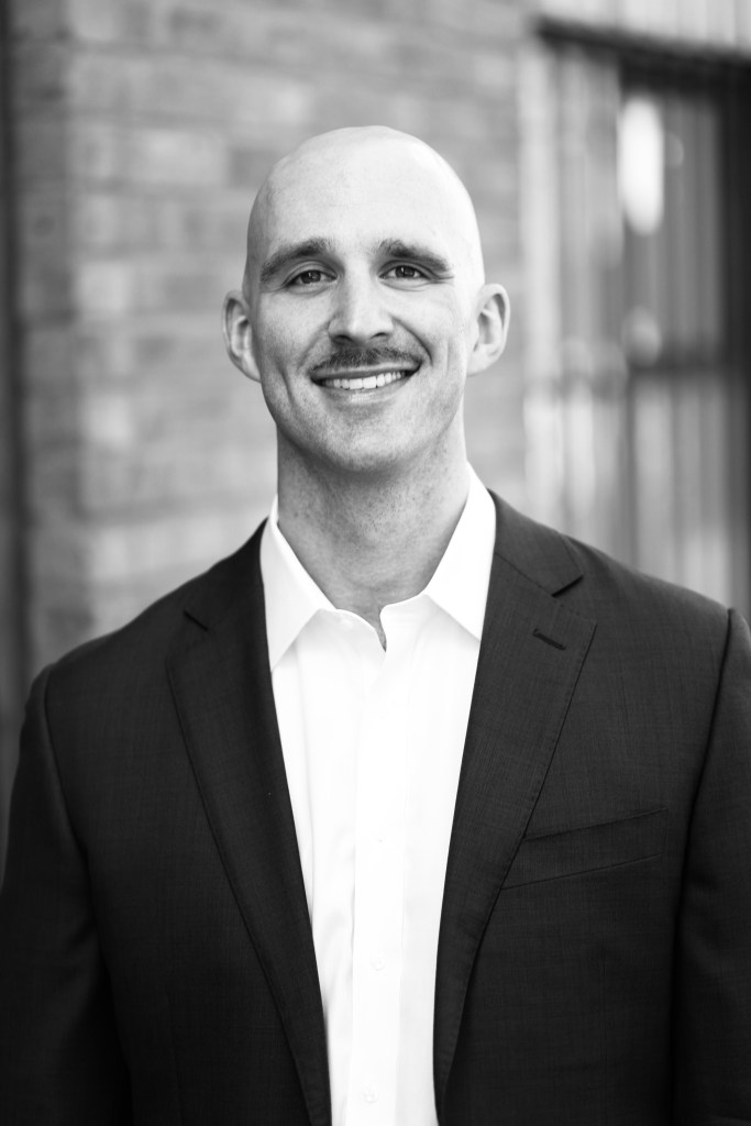 A smiling man with a bald head and mustache, wearing a dark suit jacket over a white shirt, standing in front of a brick wall.